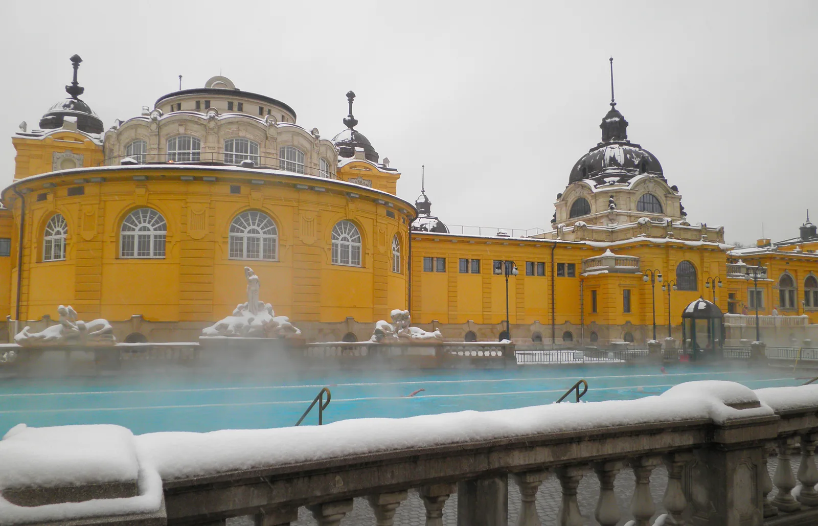 La piscina exterior de los Baños Széchenyi echando vapor en invierno, con el edificio amarillo neobarroco detrás